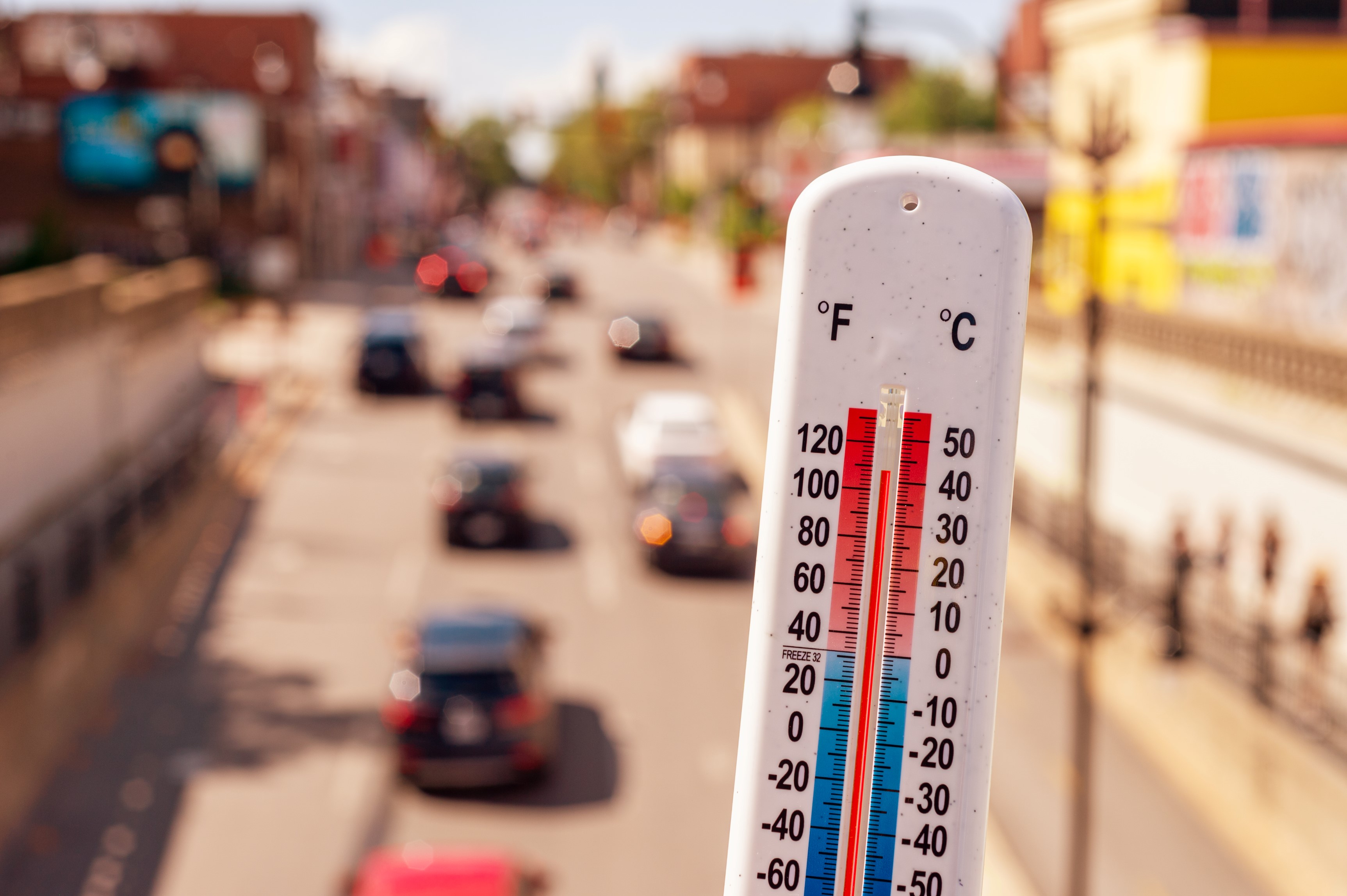 Der Betrachter blickt von erhöhter Position eine dicht befahrene Straße hinunter. Die Straße wird von vielen Häusern gesäumt und es ist ein sonniger Tag. Sehr dicht im Vordergrund ist ein Thermometer zu sehen, das 43°C anzeigt.