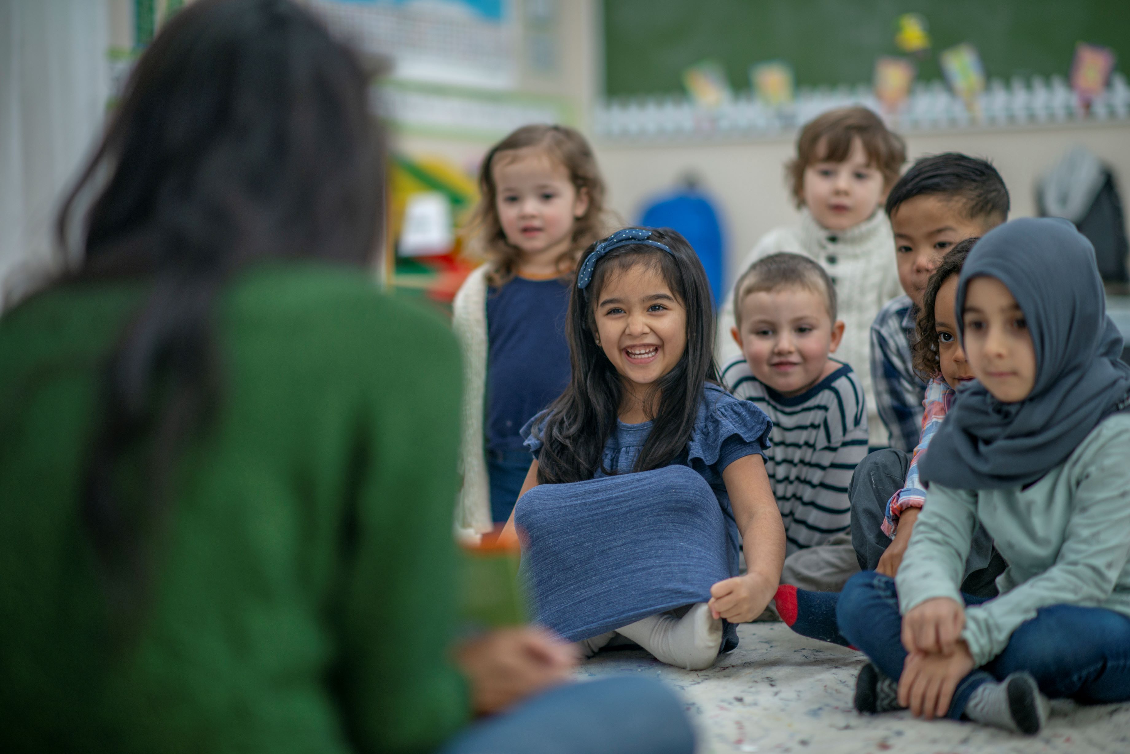 Der Betrachter befindet sich im Rücken einer jungen Lehrerin, die eine kleine Gruppe Kinder betreut. Diese knien auf dem Boden vor ihr und sind aufmerksam und fröhlich. Die meisten Kinder haben erkennbar einen Migrationshintergrund.