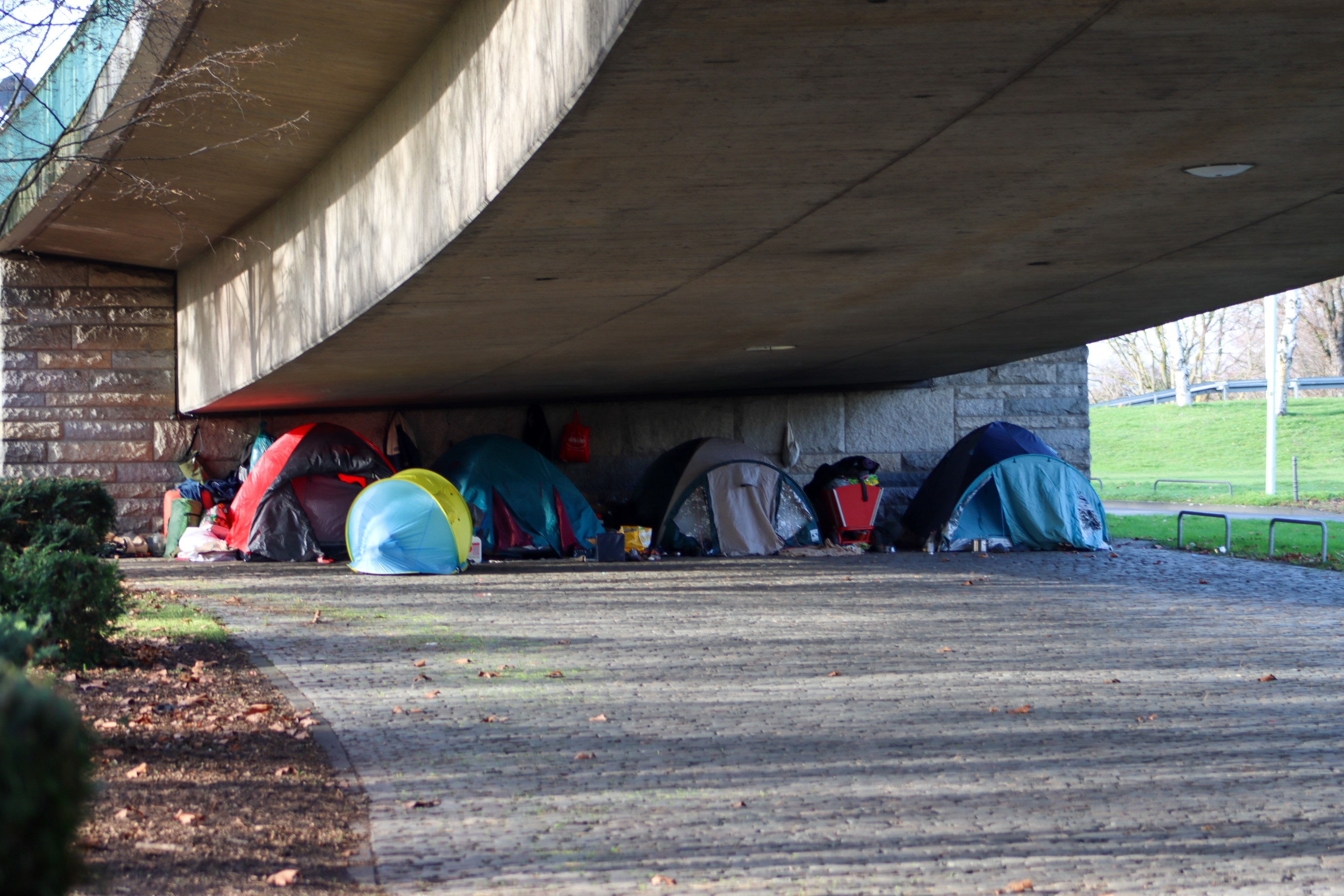 Der Betrachter steht bei sonnigem Wetter halb unter einer Autobrücke, die jedoch nicht hoch ist. Am niedrigen Fuß der Brücke sind mehrere verschiedene Zelte zu sehen, in denen offenbar Menschen wohnen.