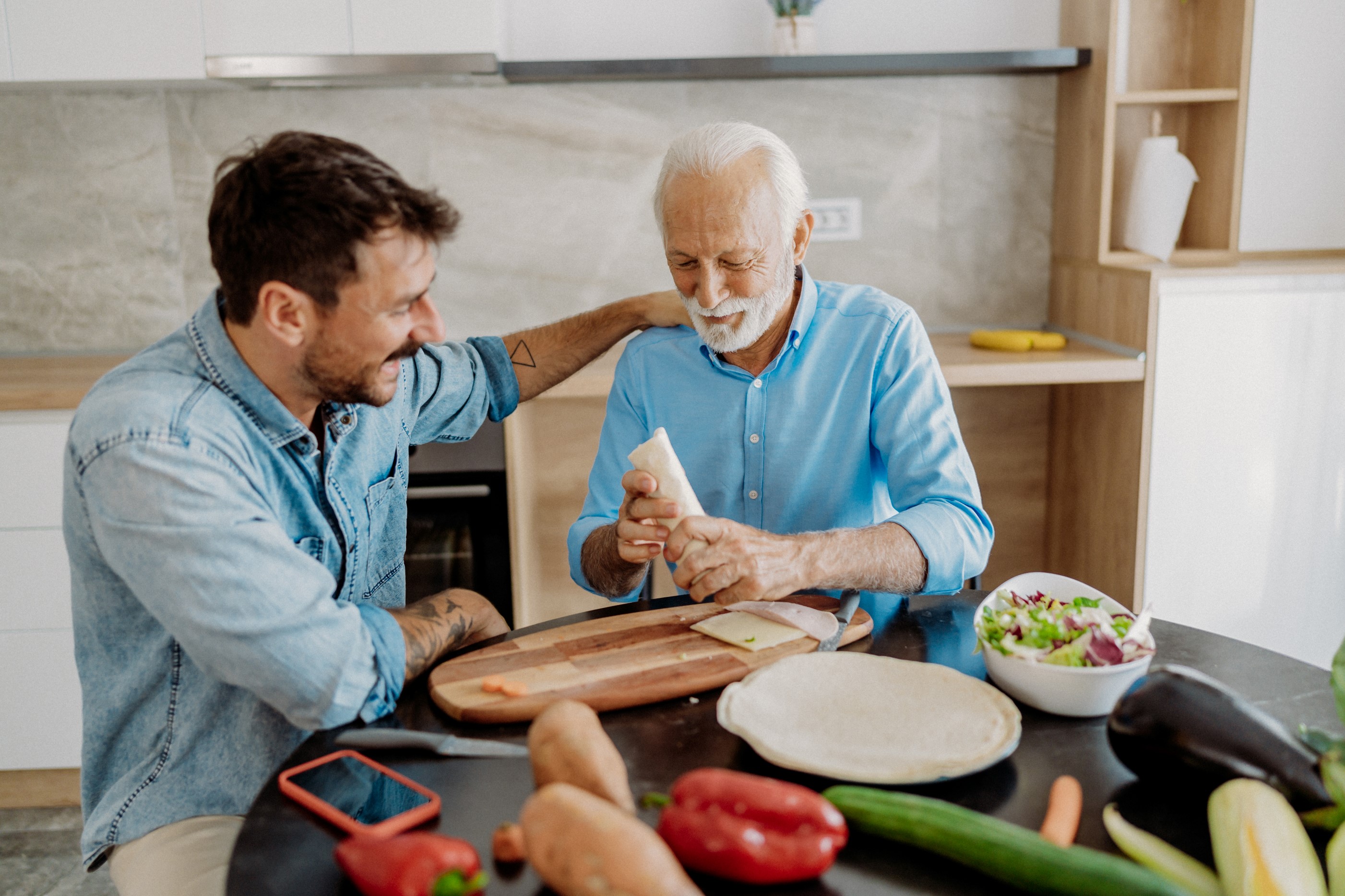 Ein alter Mann mit weißen Haaren sitzt mit einem jüngeren Mann an einem Küchentisch, auf dem Gemüse, ein Schneidbrett und ein Küchenmesser liegen. Der alte Mann hält einen Wrap in der Hand, der jüngere Mann legt ihm liebevoll eine Hand auf die Schulter.