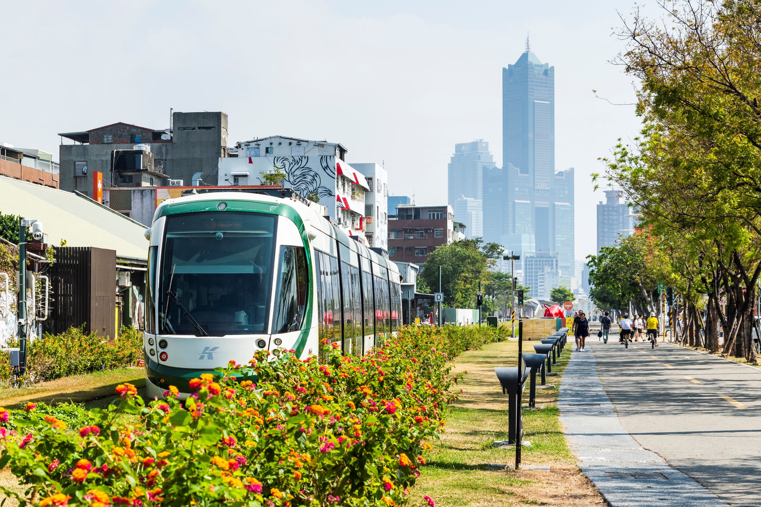 Es ist eine moderne Straßenbahn zu sehen. Die Bahntrasse ist durch ein langes Blumenbeet getrennt von einem breiten Asphaltweg auf dem Fußgänger und Radfahrer sind. Im Hintergrund sind Wolkenkratzer zu sehen.